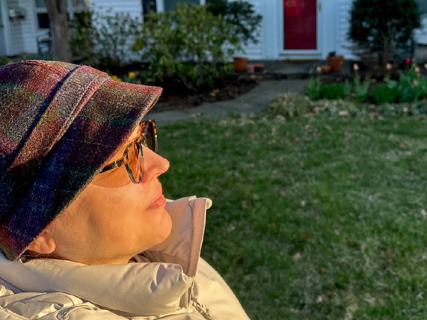 Woman in a winter hat and coat outdoors in front of a house facing the rising sun.